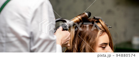 The female hairdresser is curling hair for a brown-haired young caucasian woman in a beauty salon 94880574