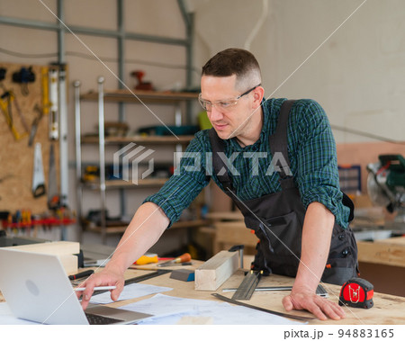 Portrait of a carpenter in protective glasses and work overalls uses a laptop in a workshop. 94883165