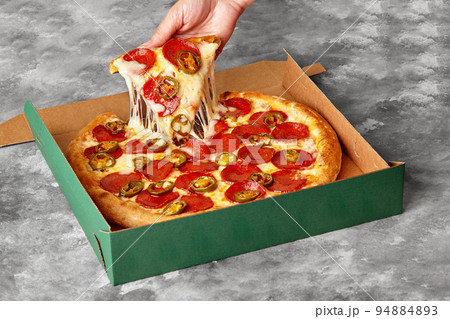 Female hand taking slice of pizza with melted cheese, pepperoni and jalapeno from cardboard box Female hand taking slice of pizza with melted cheese, pepperoni and jalapeno from cardboard box 94884893