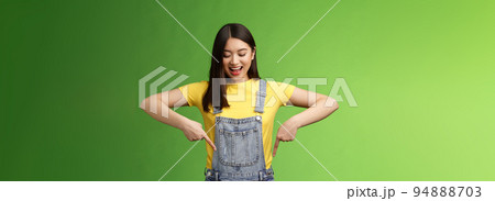 Impressed entertained cute asian girl watching pointing down interested, observe cool promo, smiling broadly, fascinated gaze bottom advertisement, take look curious thing, green background 94888703
