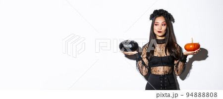 Thoughtful stylish woman in witch costume, picking pumpkin for halloween, making decision while standing over white background 94888808
