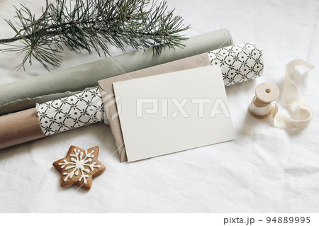Christmas stationery. Empty greeting card, invitation mockup. Gingerbread cookie, green gift wrapping papers and pine tree branches on white linen tablecloth in sunlight. Winter festive flatlay, top 94889995