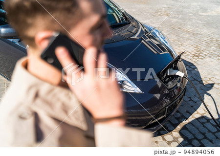 Man talks on the phone while his car is charging at the station Man talks on the phone while his car is charging at the station 94894056