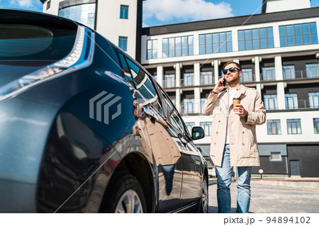 Handsome man with cup of coffee in his hand talking on the phone while standing near his car Handsome man with cup of coffee in his hand talking on the phone while standing near his car 94894102