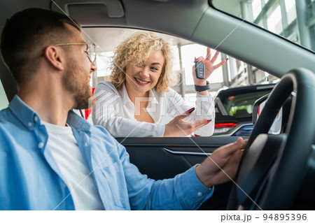 Young couple buying new car. Woman shows key to new auto to her husband who is sitting in car at dealership Young couple buying new car. Woman shows key to new auto to her husband who is sitting in car at dealership 94895396