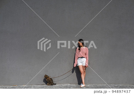 A young woman walks with a gray tabby cat on a leash against a gray wall. A young woman walks with a gray tabby cat on a leash against a gray wall. 94897437