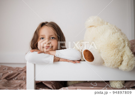Health and beauty concept - little girl with teddy bear lying on bed at home. 94897809
