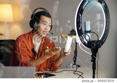 Modern young Asian male influencer sitting at desk in home studio making promotion or review video for new cleansing gel or foam Modern young Asian male influencer sitting at desk in home studio making promotion or review video for new cleansing gel or foam 94897859