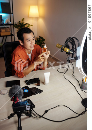 Vertical high angle view of young adult Asian male influencer making review of new beauty products demonstrating moisturizing serum or nourishing oil on camera 94897867