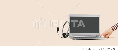 Front view of slim laptop with headset on beige. Female hand turning on. Distant learning. working from home, online courses or support. Audio podcast headphones . Helpdesk or call center banner 94899429