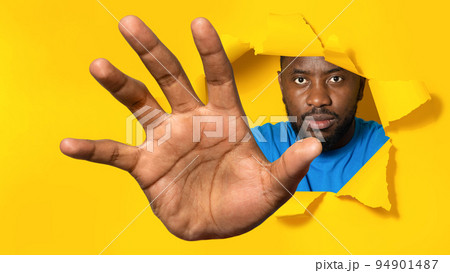 Serious african american man reaching through hole in torn yellow paper, trying to grab something Serious african american man reaching through hole in torn yellow paper, trying to grab something 94901487