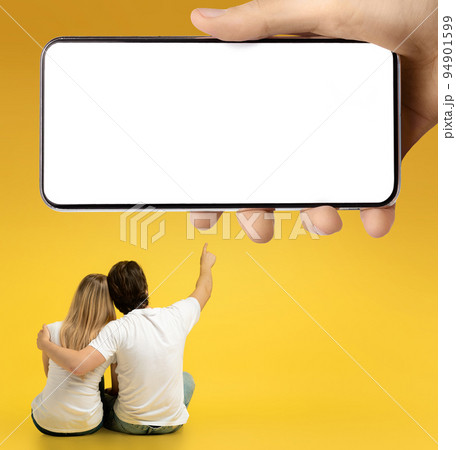 Love and romantic. Young caucasian guy hugging lady, pointing finger at huge smartphone with empty screen Love and romantic. Young caucasian guy hugging lady, pointing finger at huge smartphone with empty screen 94901599