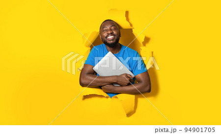 Gadget lover. Happy black man hugging laptop computer and smiling, posing in torn hole of yellow paper background 94901705