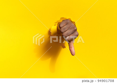 African american male hand showing thumb down through ripped hole in yellow paper background, free space 94901708