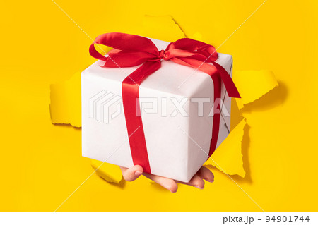 Hand giving gift box through torn yellow paper background, closeup. Special offer or sale concept 94901744