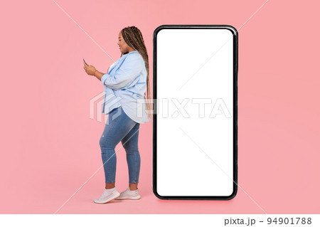 Black Female Using Phone Standing Near Large Smartphone, Pink Background Black Female Using Phone Standing Near Large Smartphone, Pink Background 94901788