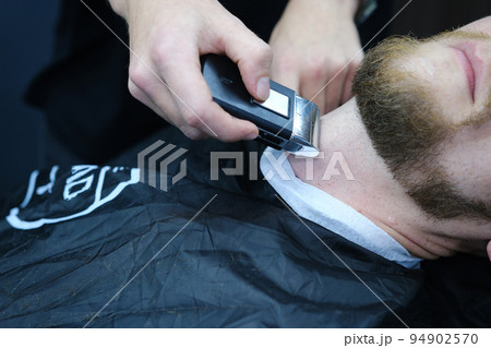 Professional Master barber shaves the client's beard with a electric trimmer. Haircut of a man's beard in a barber shop. Barber Men. Advertising and barber shop concept Professional Master barber shaves the client's beard with a electric trimmer. Haircut of a man's beard in a barber shop. Barber Men. Advertising and barber shop concept 94902570