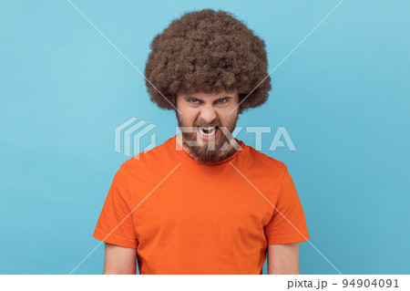 Portrait of angry man with Afro hairstyle wearing orange T-shirt looking at camera, having aggressive facial expression, expressing negative, hatred. Indoor studio shot isolated on blue background. 94904091