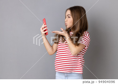 Portrait of pretty little girl wearing striped T-shirt sending kiss on smartphone camera while communicating by video call or streaming vlog. Indoor studio shot isolated on gray background. 94904096