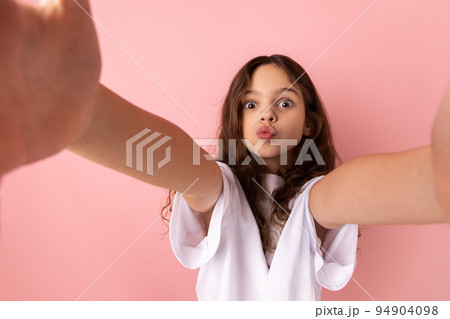 Portrait of adorable little girl wearing white T-shirt sending air kisses while taking selfie POV, flirting, expressing romantic feelings. Indoor studio shot isolated on pink background. 94904098