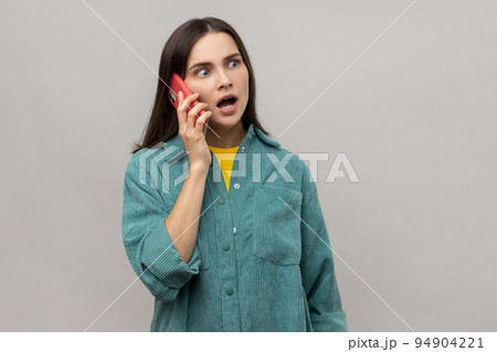 Gossip girl. Exited woman standing, using and making call on her smartphone with wondering face and talking with friend, wearing casual style jacket. Indoor studio shot isolated on gray background. Gossip girl. Exited woman standing, using and making call on her smartphone with wondering face and talking with friend, wearing casual style jacket. Indoor studio shot isolated on gray background. 94904221