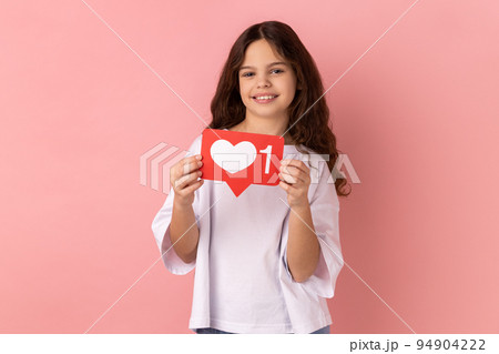 Portrait of little girl wearing white T-shirt holding social media heart Like icon and smiling to camera, recommending to follow and love content. Indoor studio shot isolated on pink background. 94904222