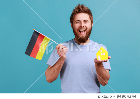 Portrait of positive optimistic handsome bearded man holding toy paper house and flag of germany in hand, moving into another country. Indoor studio shot isolated on blue background. 94904244
