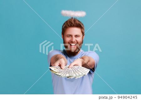 Portrait of rich handsome satisfied bearded angelic man with holy nimbus over his head holding out big fan of dollar banknotes, win the lottery. Indoor studio shot isolated on blue background. 94904245