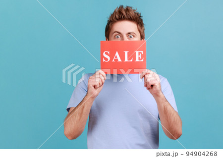 Portrait of handsome young adult man in T-shirt peeping from red card with sale inscription, mega shopping, black friday discounts. Indoor studio shot isolated on blue background. 94904268