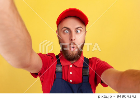 Childish playful worker man with wearing red cap and blue uniform taking selfie, looking at camera with crossed eyes POV, point of view of photo. Indoor studio shot isolated on yellow background. Childish playful worker man with wearing red cap and blue uniform taking selfie, looking at camera with crossed eyes POV, point of view of photo. Indoor studio shot isolated on yellow background. 94904342
