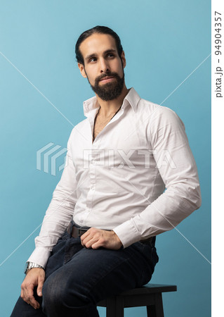Unsmiling serious dark haired attractive man with beard wearing white shirt and black trousers, looking away with attentive look while sitting on chair. Indoor studio shot isolated on blue background. Unsmiling serious dark haired attractive man with beard wearing white shirt and black trousers, looking away with attentive look while sitting on chair. Indoor studio shot isolated on blue background. 94904357