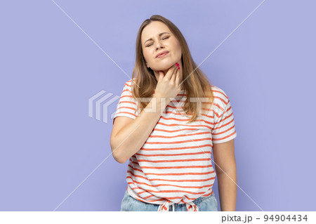 Portrait of sick blond woman wearing striped T-shirt touching her neck, suffering sore throat, viral infection or flu symptoms. Indoor studio shot isolated on purple background. 94904434