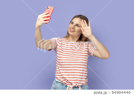 Portrait of cheerful blond woman wearing striped T-shirt with mobile phone phone and having video call, having hi or bye with hand. Indoor studio shot isolated on purple background. Portrait of cheerful blond woman wearing striped T-shirt with mobile phone phone and having video call, having hi or bye with hand. Indoor studio shot isolated on purple background. 94904440