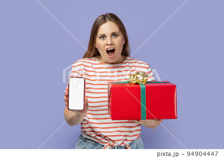 Amazed excited woman wearing striped T-shirt holding red present box and showing cell phone with white screen with copy space for advertisement. Indoor studio shot isolated on purple background. Amazed excited woman wearing striped T-shirt holding red present box and showing cell phone with white screen with copy space for advertisement. Indoor studio shot isolated on purple background. 94904447