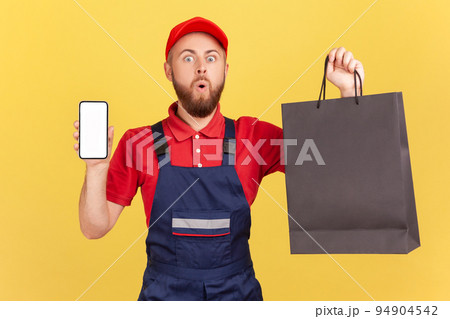 Portrait of astonished delivery man wearing blue uniform holding black shopping bag and cell phone with empty display for advertisement. Indoor studio shot isolated on yellow background. Portrait of astonished delivery man wearing blue uniform holding black shopping bag and cell phone with empty display for advertisement. Indoor studio shot isolated on yellow background. 94904542