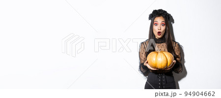 Surprised asian girl in witch costume, holding pumpkin and gasping amazed at camera, preparing for halloween holiday, standing over white background Surprised asian girl in witch costume, holding pumpkin and gasping amazed at camera, preparing for halloween holiday, standing over white background 94904662