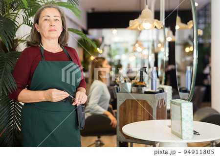 Confident elderly female hairdresser standing in hair salon 94911805