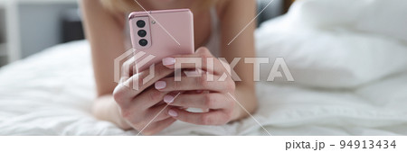 Young woman lying on bed and holding mobile phone in her hands closeup Young woman lying on bed and holding mobile phone in her hands closeup 94913434