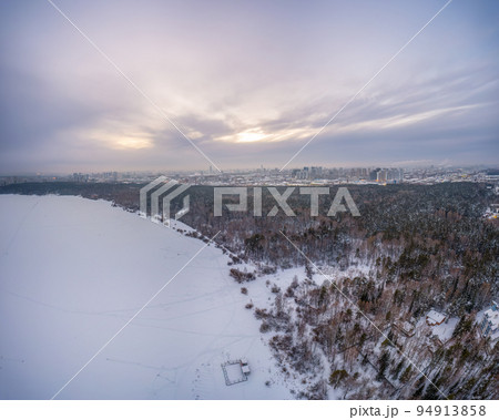 Snow-covered forest on lake shore with ice at sunset and the city on horizon, auerial view 94913858