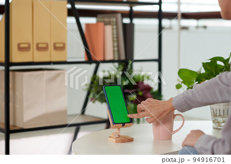 Hand of girl sitting by workplace in home office and pointing at smartphone blank green screen while starting new mobile application 94916701