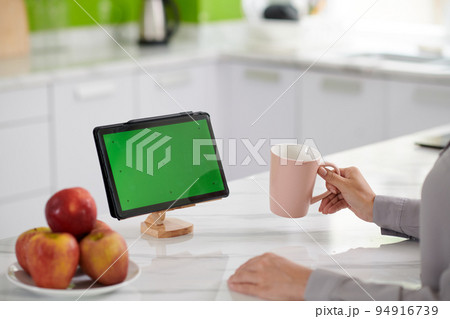 Green screen of tablet standing on wooden holder on white marble kitchen table where young home office worker sitting Green screen of tablet standing on wooden holder on white marble kitchen table where young home office worker sitting 94916739