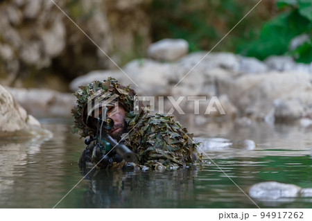 A military man or airsoft player in a camouflage suit sneaking the river and aims from a sniper rifle to the side or to target. A military man or airsoft player in a camouflage suit sneaking the river and aims from a sniper rifle to the side or to target. 94917262