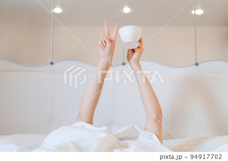 Young woman with a coffee mug in bed with white linens. Minimal happy morning Young woman with a coffee mug in bed with white linens. Minimal happy morning 94917702