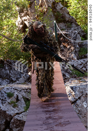 A military man or airsoft player in a camouflage suit sneaking the rope bridge and aims from a sniper rifle to the side or to target.  94918040