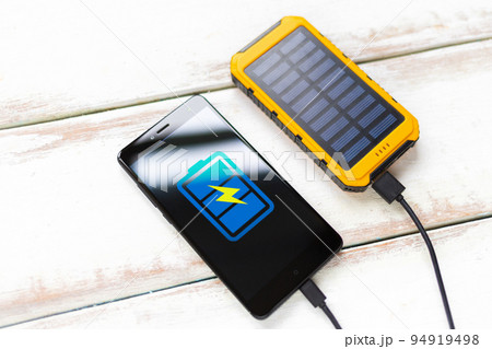 Concept of modern technologies and power bank. Smartphone with a blue battery icon, charged from a portable solar charger. White wooden background with stripes. Close up. Flat line 94919498
