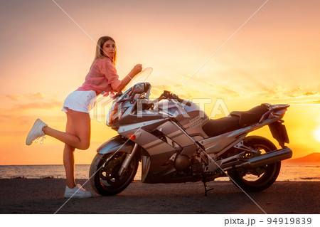 Caucasian young slim woman posing on a motorcycle. Golden sunset and ocean on the background with copy space. Freedom and mototrips Caucasian young slim woman posing on a motorcycle. Golden sunset and ocean on the background with copy space. Freedom and mototrips 94919839