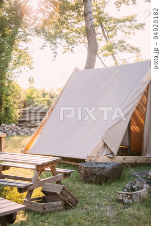 Rural camping. Rest zone. Close nature. Canvas tent with picnic desk on sunshine yard and wooden boxes outdoor. 94920182