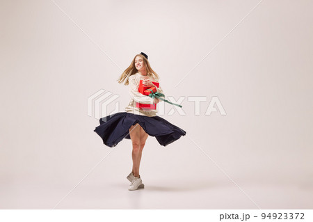 Portrait of beautiful happy girl in warm sweater and skirt dancing with bright festive gift box on gray background. New Year holidays, happiness concept 94923372