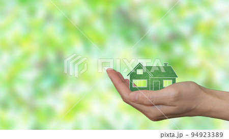 In the palm of a woman's hand, an eco-green house. Blurry green leaves in the background In the palm of a woman's hand, an eco-green house. Blurry green leaves in the background 94923389