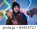 Authentic young woman in fur coat with leopard pattern and black hat standing on background of festive Christmas lights, holiday decorations. Shallow depth of field, selective soft focus on foreground 94924727